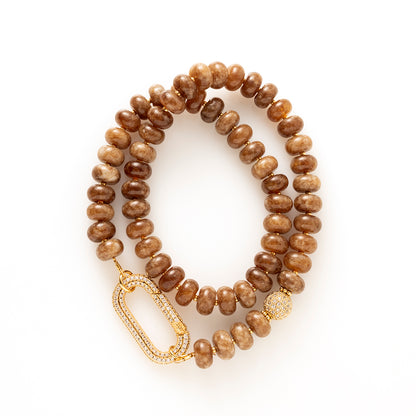 Earth Essence – Signature Clasp is uitgevoerd met dezelfde warme bruine natuursteen kralen. De tint is diep en natuurlijk, waardoor de ketting moeiteloos combineert met zowel lichte als donkere outfits.

Het verschil zit in het zirkonia slot. Deze variant heeft een ander type sluiting (veersluiting) dat een eigen uitstraling geeft: groot, sprankelend en opvallend als designaccent. Het slot fungeert niet alleen als sluiting, maar ook als stijlvol detail dat gezien mag worden.
