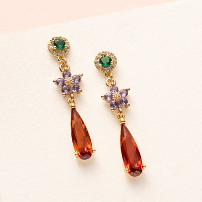 Elegant en speels: Autumn Glow – Flower begint met een groene zirkonia oorsteker, gevolgd door een bloemvormig paars tussenstuk en eindigt in een opvallende oranje druppel van crystal glas. Subtiel van boven, sprankelend van onderen – perfect voor kantoor of een etentje.