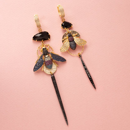 Asymmetrische statement oorbellen met grote zwarte kristalglasstenen, kleurrijke vliegen in donkerblauw, turquoise, magenta en kristal, bezaaid met zirkonia stenen. Gunmetal afwerking voor een stoere, gedurfde look. Gemaakt van 18kt gold plated brass, nikkelvrij, ideaal voor de vrouw die opvalt. Oorbellen met sluitbare ringen.